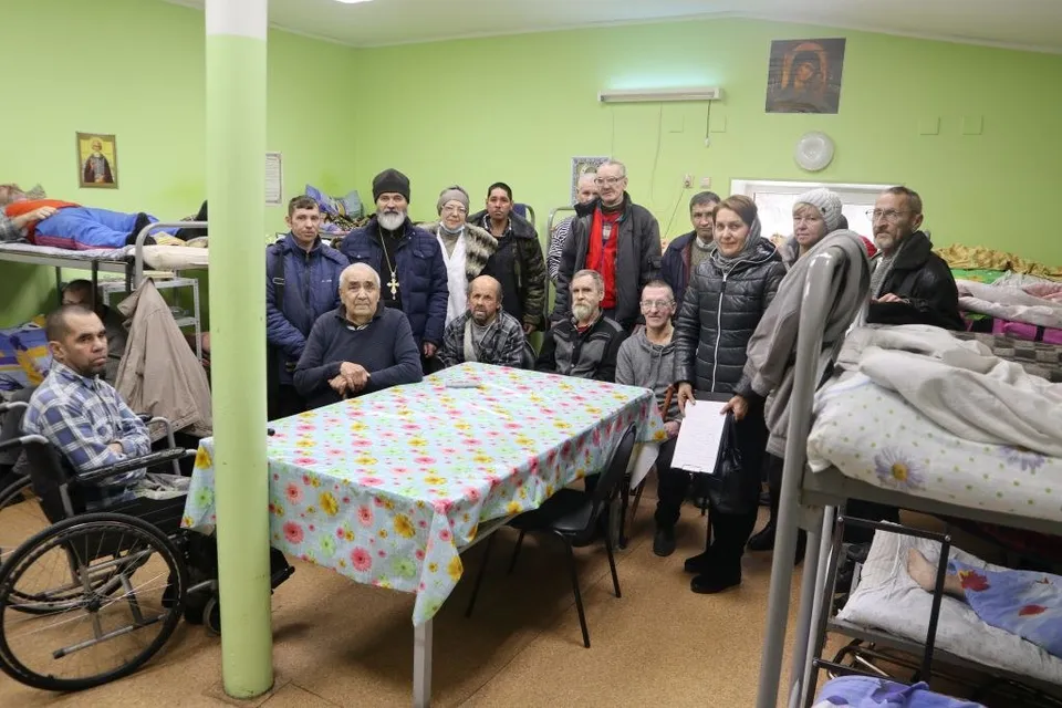 Группа мужчин, в том числе в инвалидной коляске, и священник у стола в комнате приюта с двухъярусными кроватями